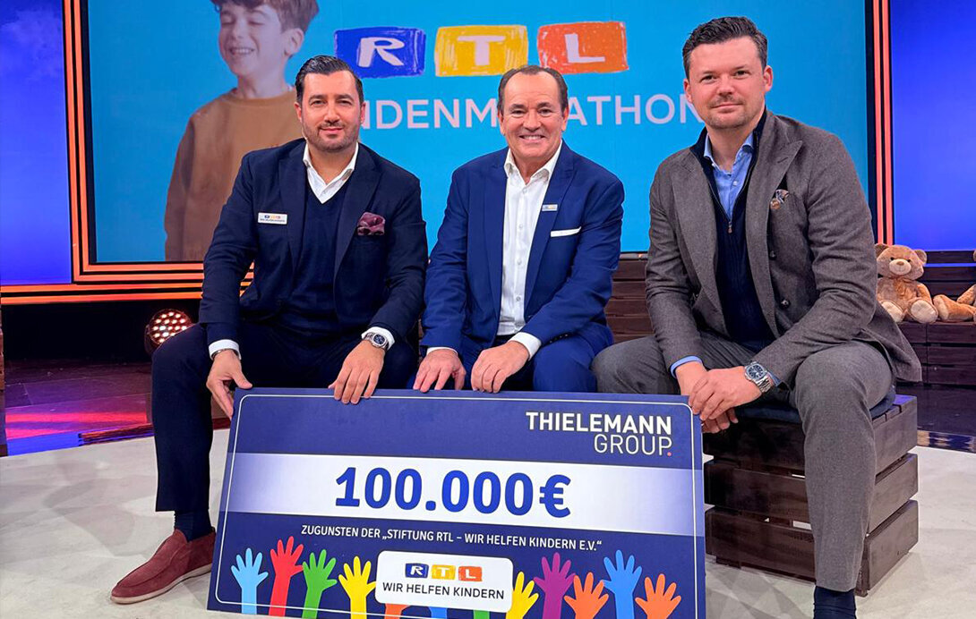 Navid Thieleman, Wolfgang Kons und Christian Flick (von links nach rechts) sitzend, davor der symbolische Scheck von 100.000 Euro